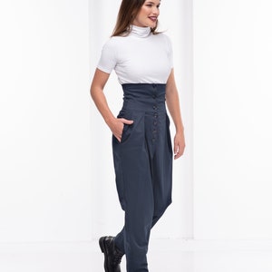 Pu&ograve; includere: Una donna indossa un top bianco a maniche corte con collo alto e pantaloni blu navy a vita alta con bottoni. I pantaloni hanno una fascia in vita ampia e una vestibilit&agrave; morbida. Scarpe in pelle nera completano l'outfit.