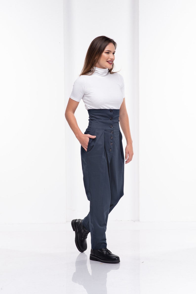 Pu&ograve; includere: Una donna indossa un top bianco a maniche corte con collo alto e pantaloni blu navy a vita alta con bottoni. I pantaloni hanno una fascia in vita ampia e una vestibilit&agrave; morbida. Scarpe in pelle nera completano l'outfit.