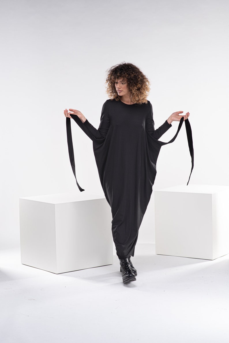 May include: A woman wearing a black long-sleeved dress with a loose, flowing silhouette. The dress has long ties that can be tied at the waist. She is standing on a white platform with her feet in black boots.