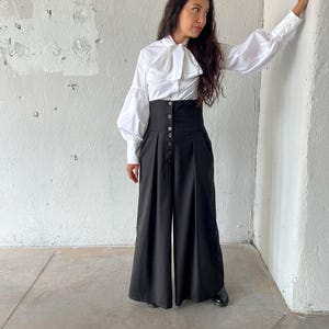 May include: A white blouse with a large bow and long, puffy sleeves paired with high-waisted, wide-leg black pants. The pants have a button-up closure. The outfit is a modern take on a classic style.