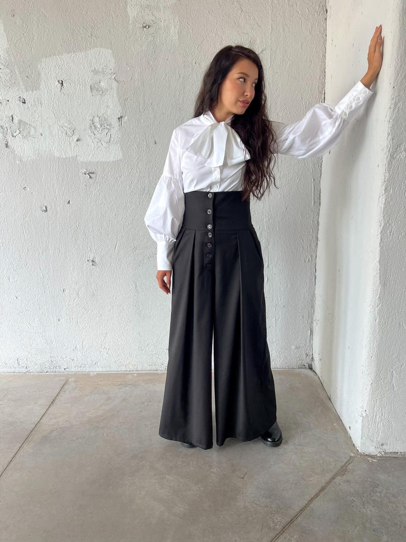 May include: A white blouse with a large bow and long, puffy sleeves paired with high-waisted, wide-leg black pants. The pants have a button-up closure. The outfit is a modern take on a classic style.