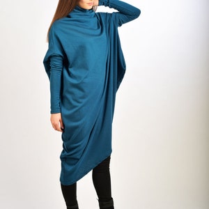May include: A teal blue long-sleeved tunic dress with a cowl neck and a draped design. The dress is worn with black leggings and black wedge heeled boots.
