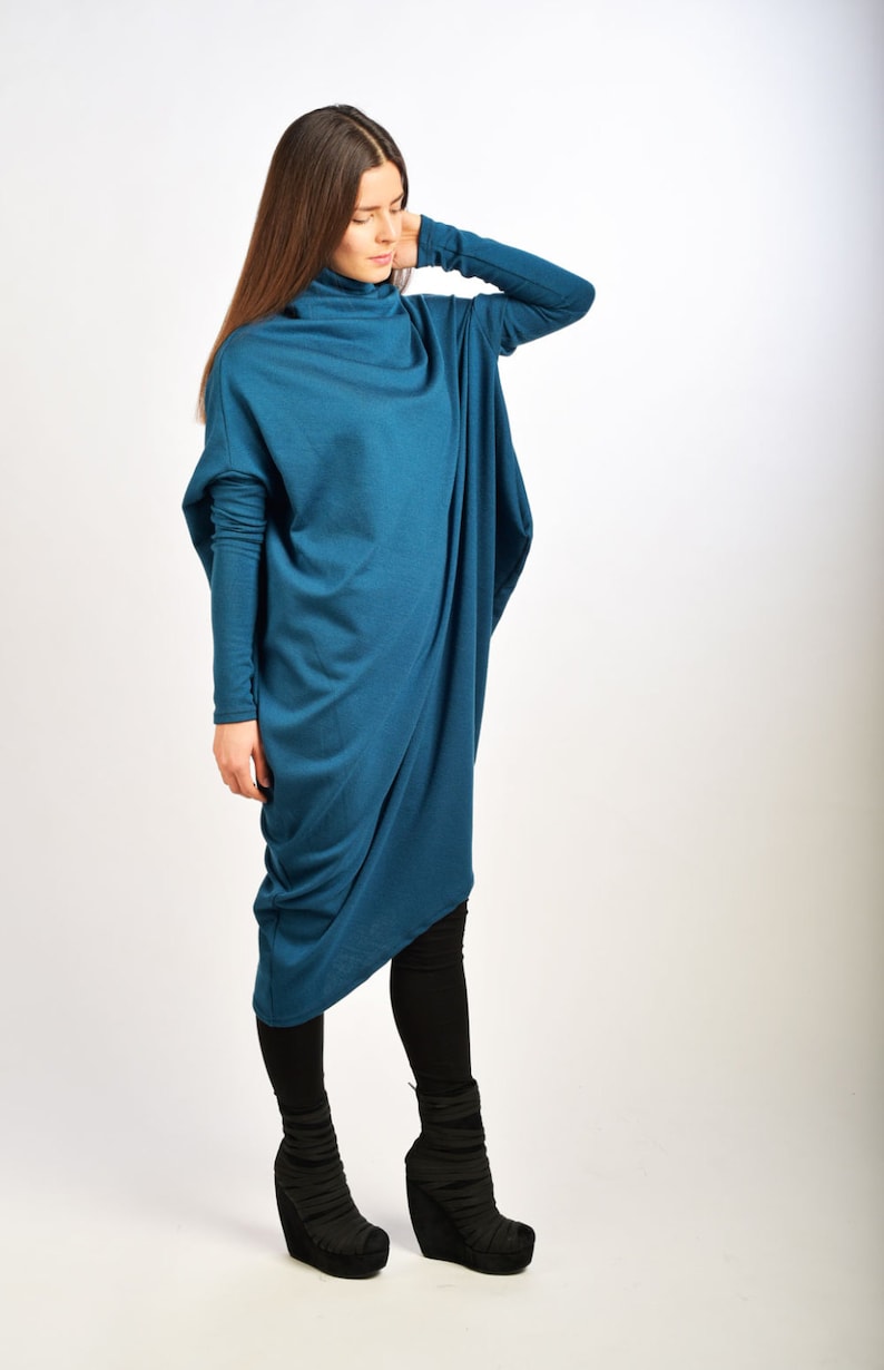 May include: A teal blue long-sleeved tunic dress with a cowl neck and a draped design. The dress is worn with black leggings and black wedge heeled boots.