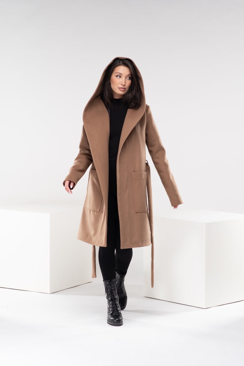 May include: A brown hooded coat with a tie belt. The coat is worn by a person with long dark hair. The person is standing on a white surface.