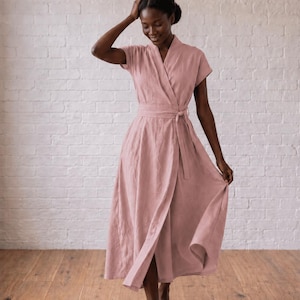 May include: A dusty rose wrap dress featuring short sleeves, a V-neckline, and a matching fabric tie belt at the waist. The dress has a midi-length skirt with a flared silhouette and is constructed from a textured fabric with visible weave details.