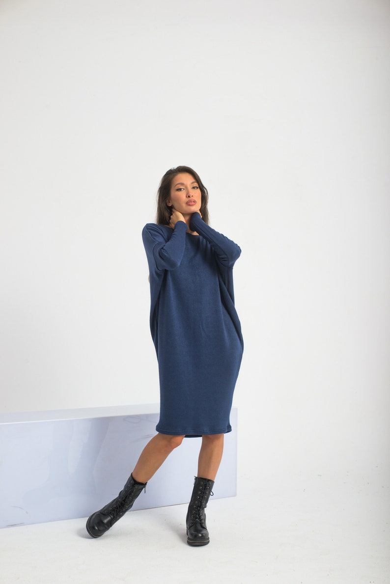 May include: A navy blue, long-sleeved dress with a relaxed fit. The dress falls to the mid-calf and is paired with black lace-up boots. The model is standing in front of a white background.