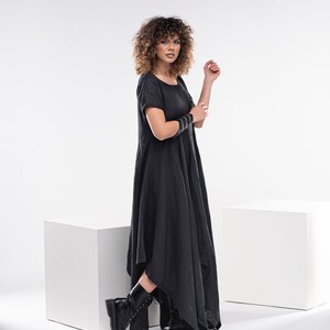 May include: A woman wearing a black, asymmetrical linen dress with short sleeves and black boots. She is standing in front of a white background.