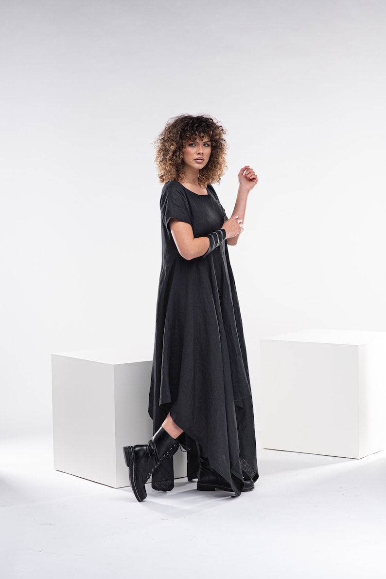 May include: A woman wearing a black, asymmetrical linen dress with short sleeves and black boots. She is standing in front of a white background.