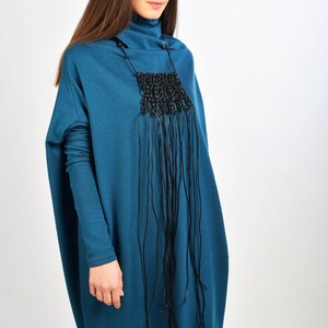 May include: A teal blue long-sleeved dress with a black fringe necklace. The dress has a loose, oversized fit.
