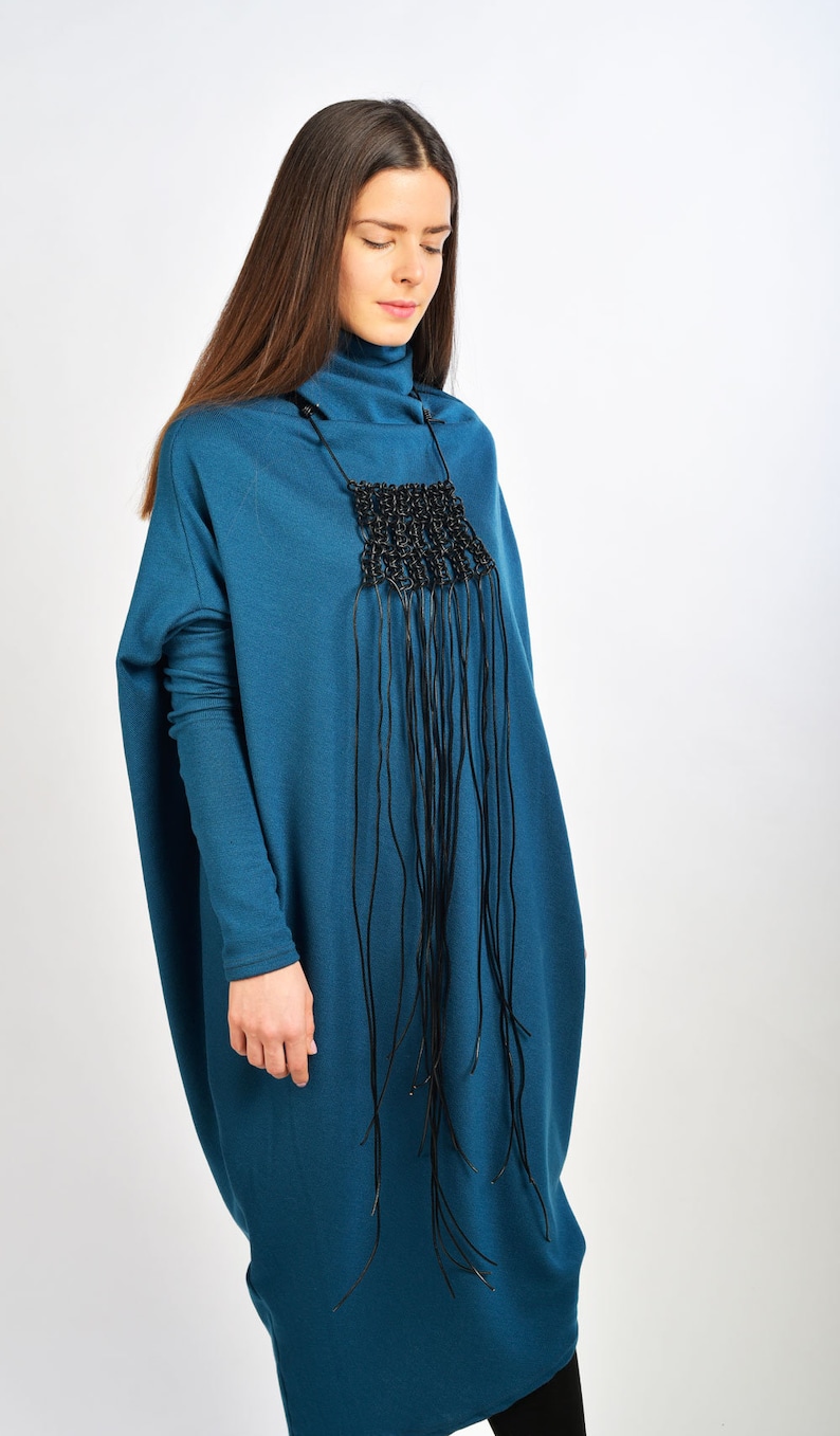 May include: A teal blue long-sleeved dress with a black fringe necklace. The dress has a loose, oversized fit.