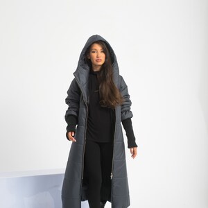 May include: A long, dark gray puffer coat with a hood and a zipper closure. The coat has a quilted design and is worn by a person with long brown hair.