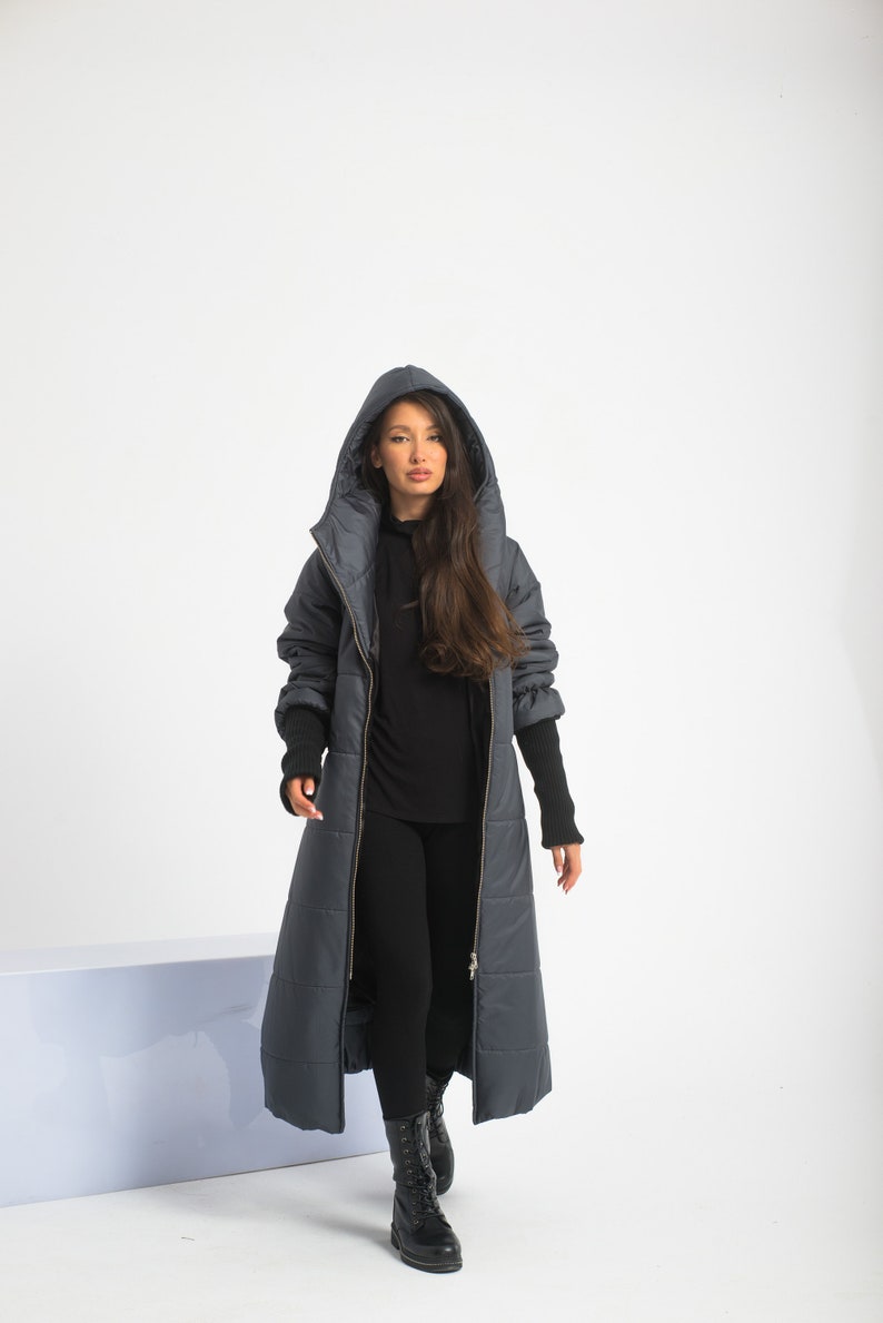 May include: A long, dark gray puffer coat with a hood and a zipper closure. The coat has a quilted design and is worn by a person with long brown hair.