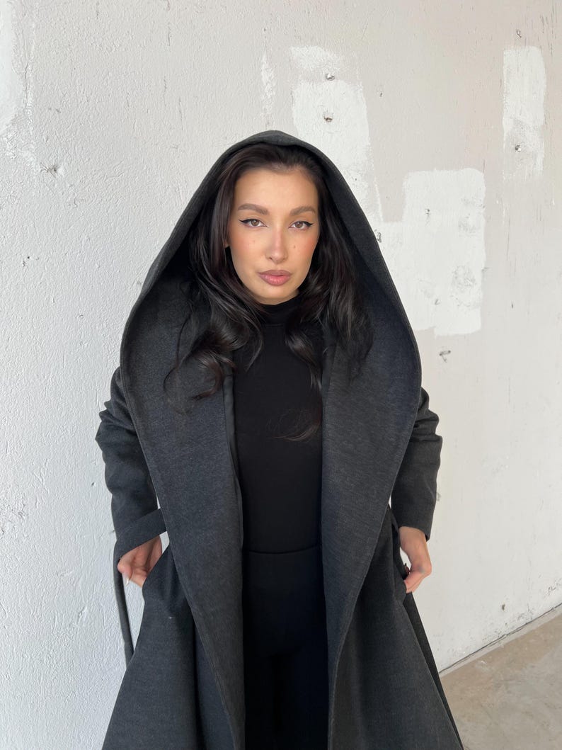 Puede incluir: Una larga gabardina gris oscuro con capucha, cuello ancho y cintur&oacute;n de anudar. La gabardina est&aacute; hecha de una tela suave y c&oacute;moda y tiene un corte holgado.