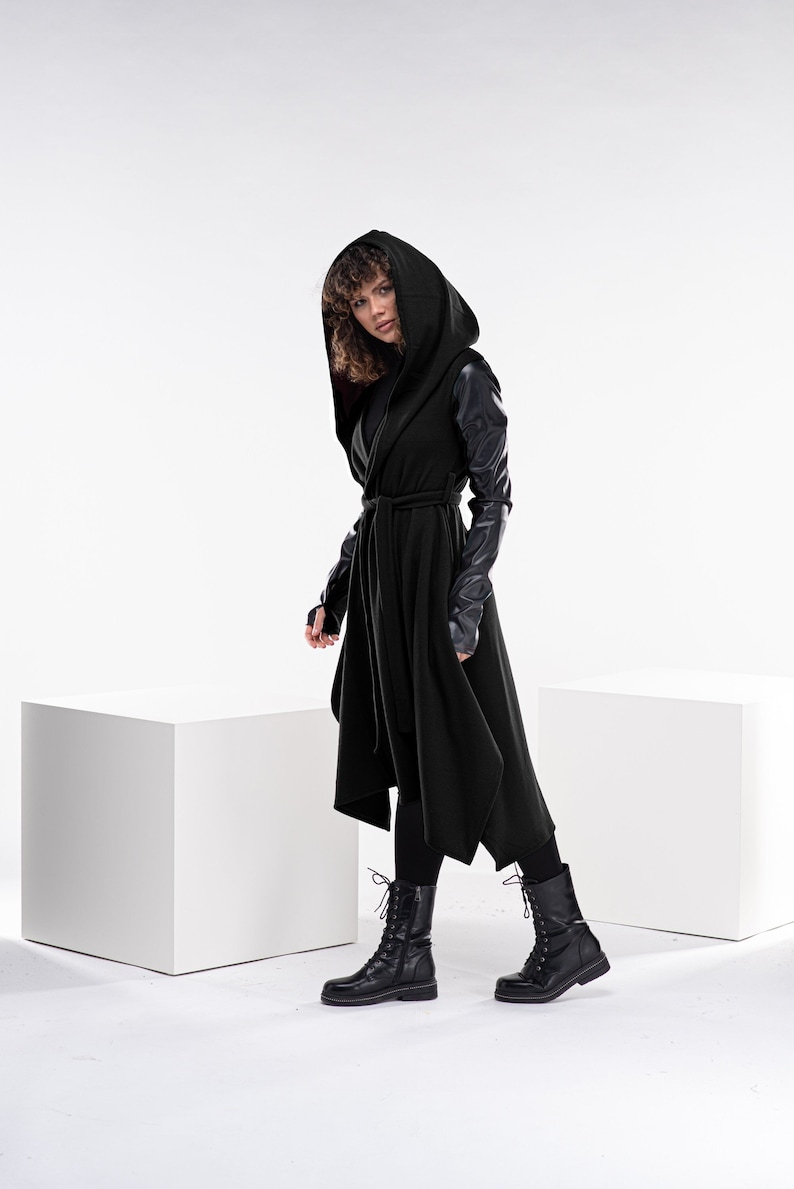 May include: A black hooded wrap coat with leather sleeves. The coat has a tie belt and a long, asymmetrical hemline. The model is wearing black boots.