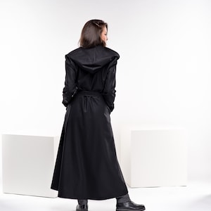 May include: A long black hooded coat with a belt. The coat is worn by a person standing on a white surface.