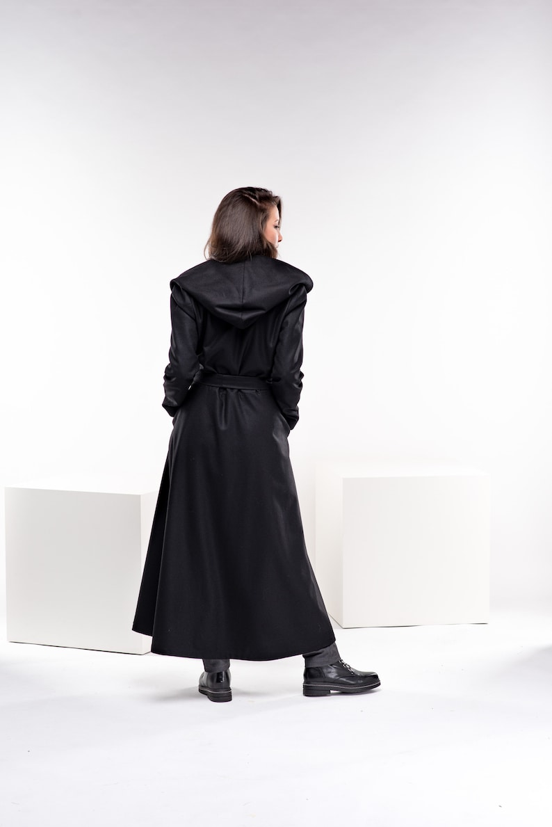 May include: A long black hooded coat with a belt. The coat is worn by a person standing on a white surface.