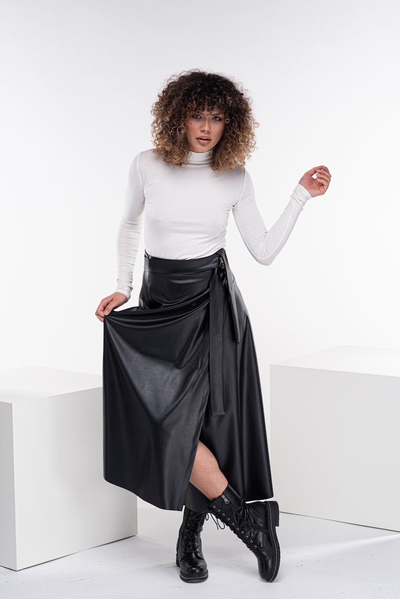 May include: A woman wearing a white turtleneck top and a black faux leather wrap skirt. She is wearing black lace-up boots and has curly brown hair.