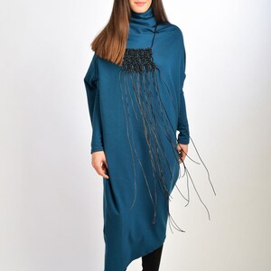 May include: A teal blue long-sleeved dress with a black fringe necklace. The dress has a loose fit and a high neckline. The model is wearing black boots.
