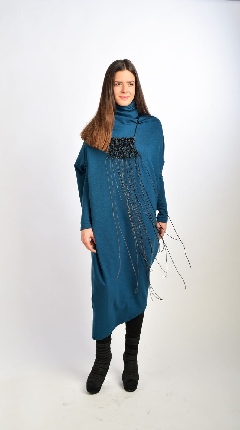 May include: A teal blue long-sleeved dress with a black fringe necklace. The dress has a loose fit and a high neckline. The model is wearing black boots.