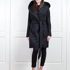 K&ouml;nnte beinhalten: Ein schwarzer Mantel mit Kapuze, passendem G&uuml;rtel und Taschen. Der Mantel wird &uuml;ber einer schwarzen Hose und metallisch-silbernen Schuhen getragen. Der Mantel hat eine lange L&auml;nge und eine breite Kapuze.