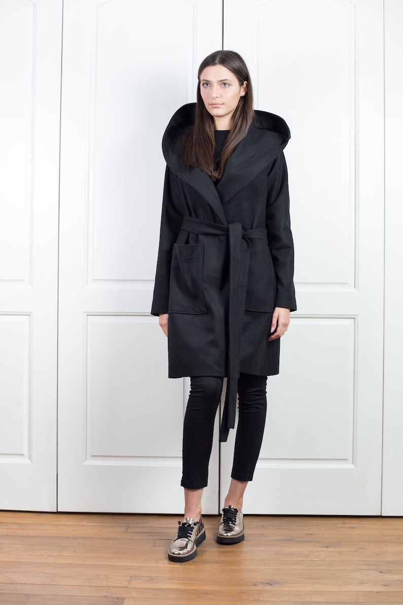 K&ouml;nnte beinhalten: Ein schwarzer Mantel mit Kapuze, passendem G&uuml;rtel und Taschen. Der Mantel wird &uuml;ber einer schwarzen Hose und metallisch-silbernen Schuhen getragen. Der Mantel hat eine lange L&auml;nge und eine breite Kapuze.