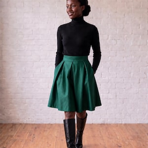 Peut inclure: Un pull à col roulé noir à manches longues associé à une jupe plissée vert forêt arrivant au genou. La jupe présente une taille haute et une silhouette évasée. La tenue est complétée par des bottes hautes en cuir noir.