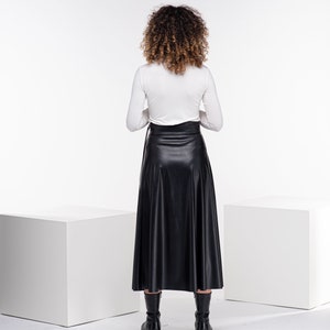 May include: A woman wearing a white long-sleeved shirt and a black faux leather skirt. She is standing in front of a white background.