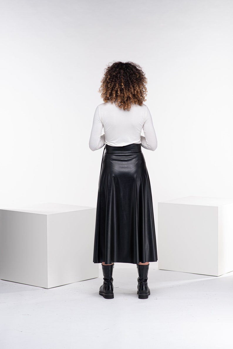 May include: A woman wearing a white long-sleeved shirt and a black faux leather skirt. She is standing in front of a white background.