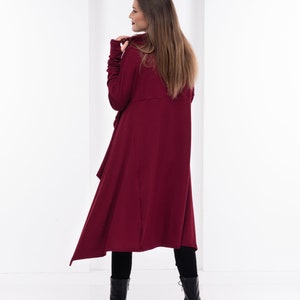 May include: A long, burgundy, asymmetrical tunic with a high neck and long sleeves. The tunic is made of a soft, stretchy fabric and has a relaxed fit. The model is wearing black leggings and black boots.