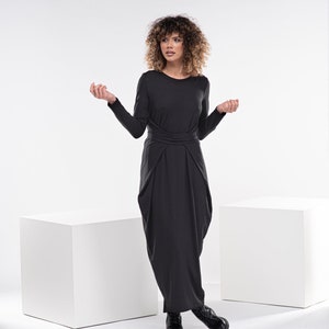 May include: A woman wearing a black long-sleeved dress with a gathered skirt and a knotted waist. She is standing on a white platform with her arms outstretched.
