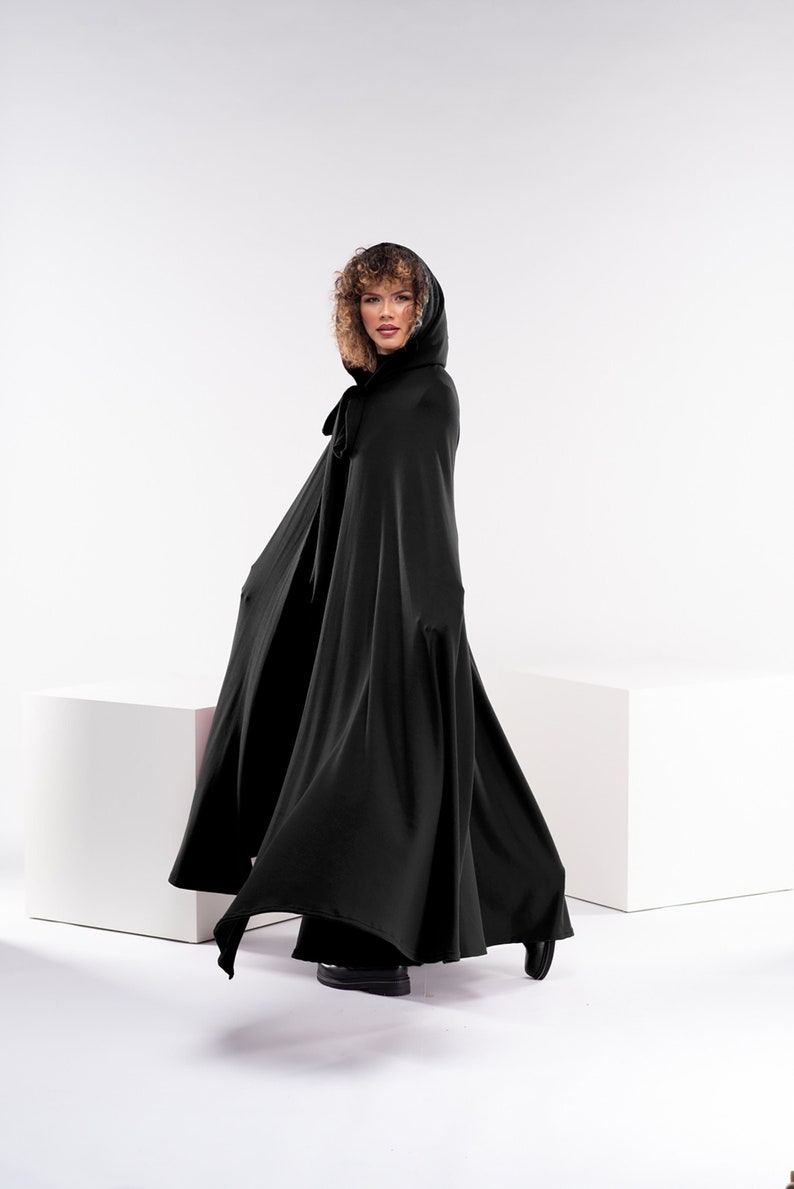 May include: A black hooded cape with a fur trim around the hood. The cape is flowing in the wind and the model is looking over her shoulder.