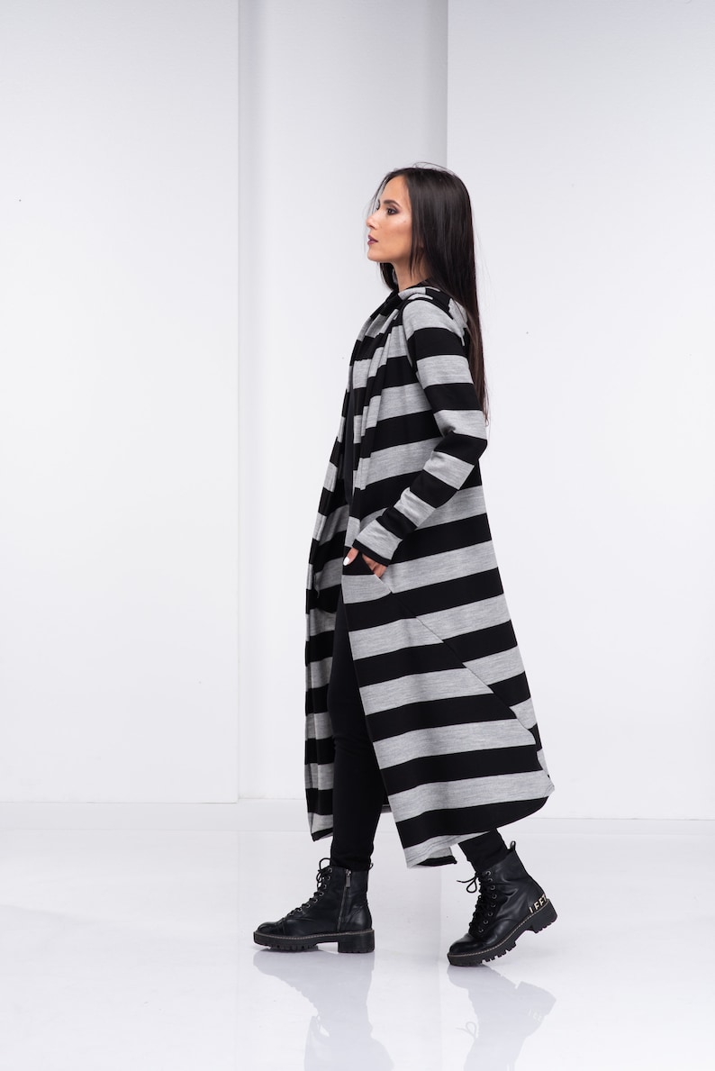 Pu&ograve; includere: Un lungo cardigan a righe nere e grigie con cappuccio. Il cardigan &egrave; aperto davanti e ha tasche sui lati. La modella indossa pantaloni neri e stivali neri.