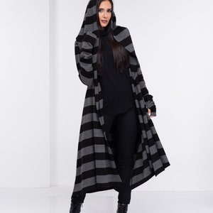 Op de afbeelding: Lange cardigan met capuchon in een zwart-grijze horizontale streep. De cardigan heeft lange mouwen en een ontwerp dat tot op de grond komt. De persoon op de foto draagt een zwarte broek en laarzen, en een zwarte top.