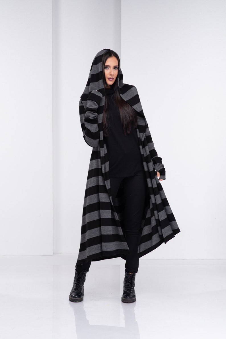 Op de afbeelding: Lange cardigan met capuchon in een zwart-grijze horizontale streep. De cardigan heeft lange mouwen en een ontwerp dat tot op de grond komt. De persoon op de foto draagt een zwarte broek en laarzen, en een zwarte top.