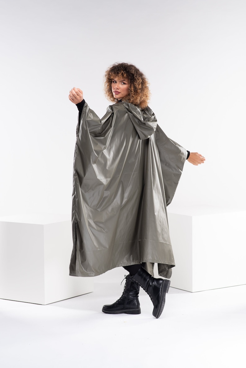 May include: A woman wearing a hooded, silver-grey raincoat with a long, flowing design. She is standing on a white surface and wearing black boots.