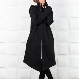 May include: A long black hooded sweatshirt with a zipper down the front. The sweatshirt has a high neck and long sleeves. The model is wearing black pants and gold shoes.
