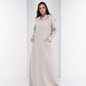 May include: A long, beige linen dress with a hood and pockets. The dress is loose-fitting and has a relaxed silhouette.
