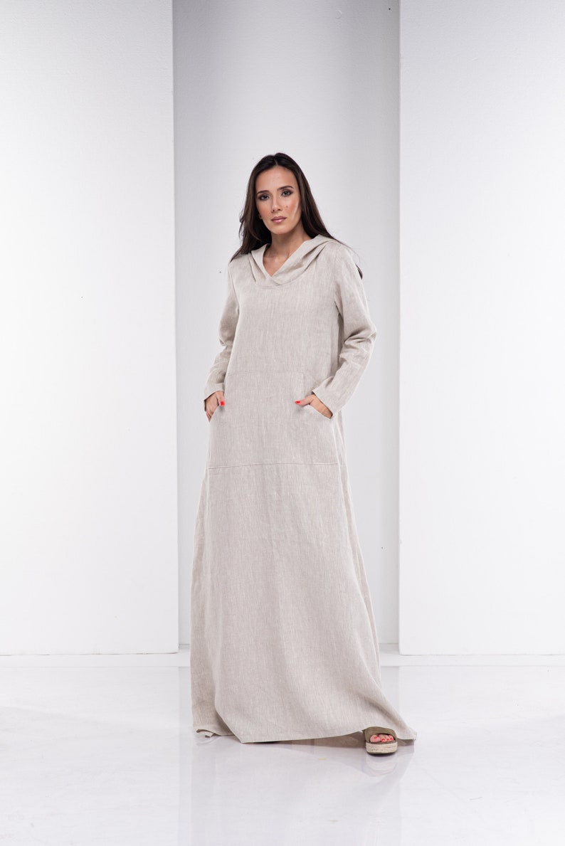 May include: A long, beige linen dress with a hood and pockets. The dress is loose-fitting and has a relaxed silhouette.