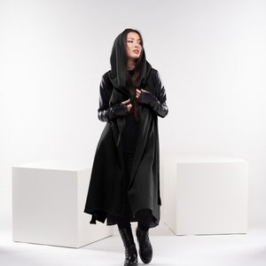 May include: A woman wearing a long, dark green hooded coat with black leather sleeves. She is standing in front of a white background.