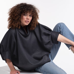 Puede incluir: Una mujer con una camisa negra de cuello alto y de corte holgado y unos vaqueros azules. Est&aacute; sentada con una pierna doblada y el pie apoyado en una superficie blanca.