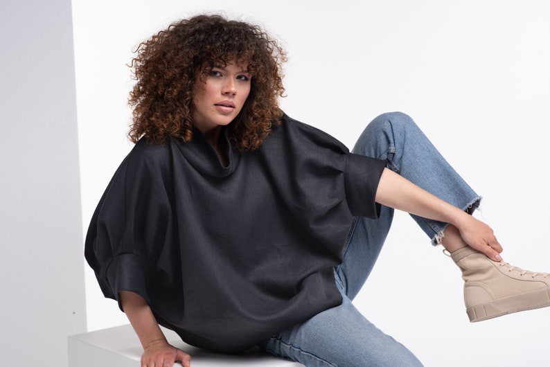 Puede incluir: Una mujer con una camisa negra de cuello alto y de corte holgado y unos vaqueros azules. Est&aacute; sentada con una pierna doblada y el pie apoyado en una superficie blanca.