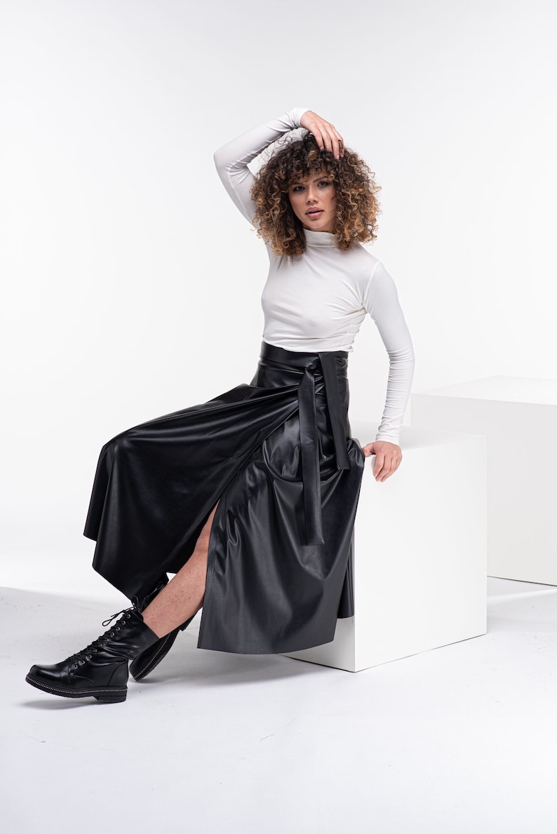 May include: A woman wearing a white turtleneck shirt and a black leather wrap skirt. She is sitting on a white cube and wearing black combat boots.