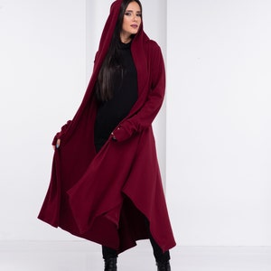 May include: A long, burgundy hooded cardigan with a draped design. The cardigan has long sleeves and a high neckline. The model is wearing black pants and black boots.