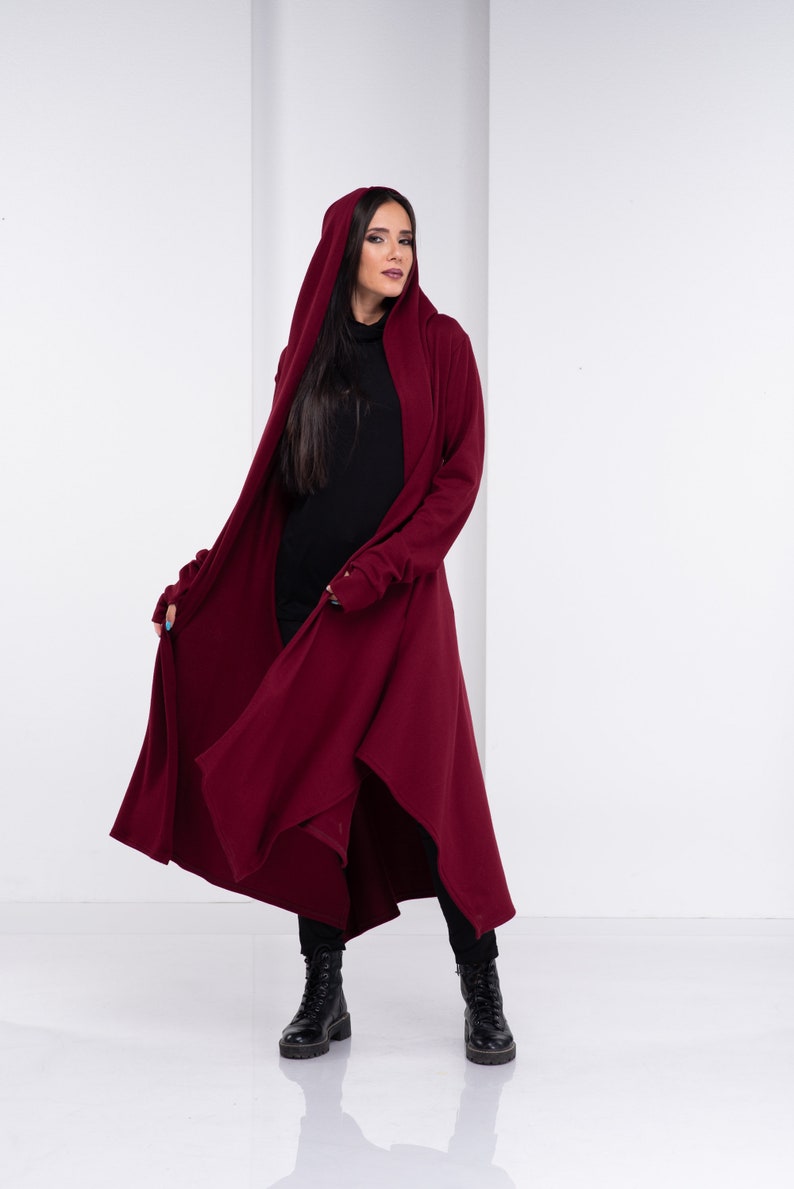 May include: A long, burgundy hooded cardigan with a draped design. The cardigan has long sleeves and a high neckline. The model is wearing black pants and black boots.