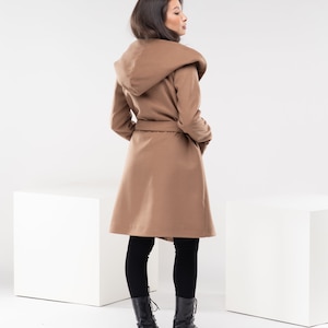 May include: A brown hooded coat with a belt, worn by a person with long dark hair. The coat is belted at the waist and has a large hood.