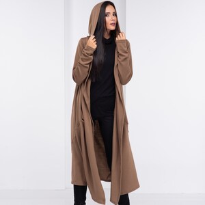 Pu&ograve; includere: Un lungo cardigan marrone con cappuccio, dalla vestibilit&agrave; rilassata e con tasche. Il cardigan &egrave; indossato sopra un maglione nero a collo alto e pantaloni neri.