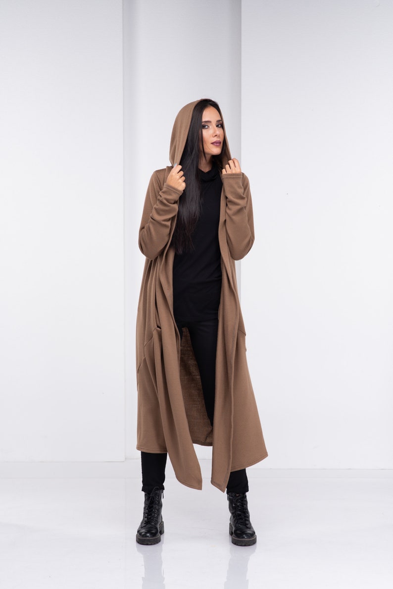 Pu&ograve; includere: Un lungo cardigan marrone con cappuccio, dalla vestibilit&agrave; rilassata e con tasche. Il cardigan &egrave; indossato sopra un maglione nero a collo alto e pantaloni neri.