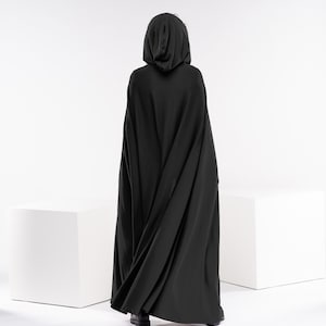 May include: A person wearing a black hooded cloak, standing in front of a white background.