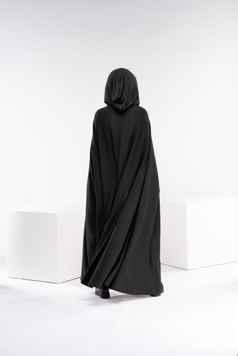 May include: A person wearing a black hooded cloak, standing in front of a white background.