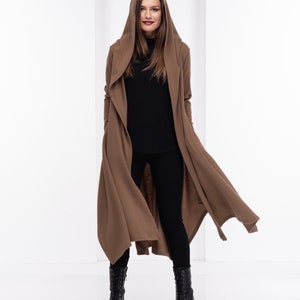 May include: A brown hooded cardigan with a long, flowing design. The cardigan is worn over a black turtleneck and black trousers. The person is wearing black boots.